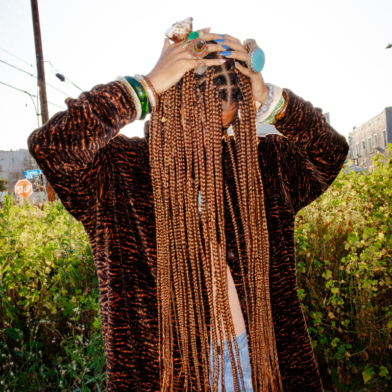 Woman outdoors wearing long Blhair™ copper-toned braids with layered jewelry and textured protective hairstyle.